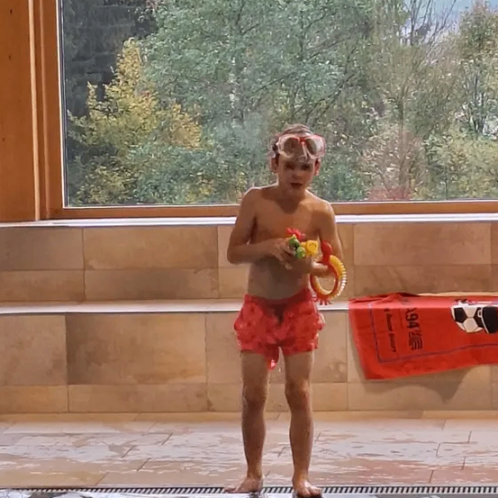 Ein junger Junge steht in einem Pool, trägt eine Schwimmbrille und hält Spielzeug. Er trägt rote Shorts. Hinter ihm zeigt ein Fenster Bäume und eine Landschaft.