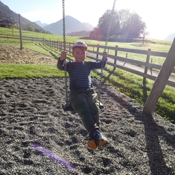 Ein Junge sitzt auf einer Schaukel in einem Spielplatz mit Schotter unter den Füßen. Hinter ihm steht ein Holzzaun, und in der Ferne befindet sich eine Grasfläche mit Bergen.