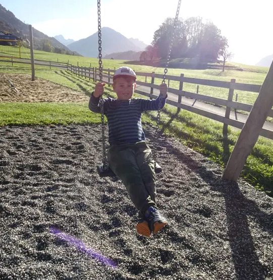 Ein Junge sitzt auf einer Schaukel in einem Spielplatz mit Schotter unter den Füßen. Hinter ihm steht ein Holzzaun, und in der Ferne befindet sich eine Grasfläche mit Bergen.