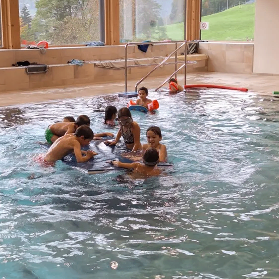 Eine Gruppe von Kindern schwimmt in einem Pool, einige tragen Schwimmbrillen und Schwimmwesten. Sie werden von einer erwachsenen Frau in der Mitte begleitet. Draußen sind durch die Glasfenster ein grasiger Hügel und Bäume zu sehen.