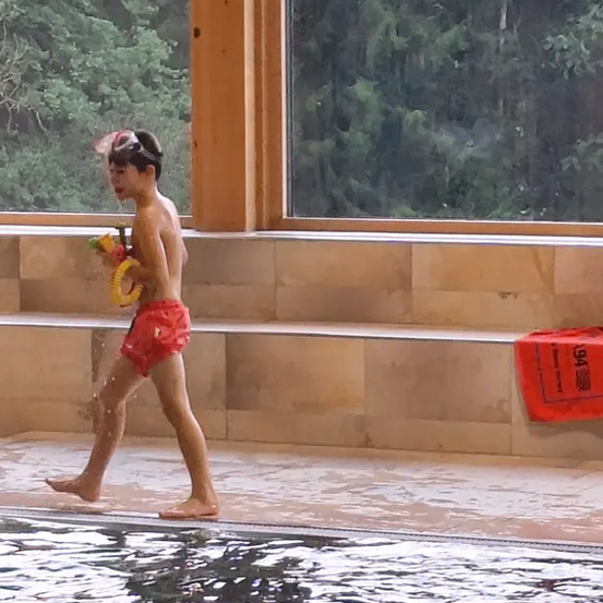 Ein Junge geht in einem Pool mit Schwimmbrille und hält ein Wasserspielzeug. Er spritzt Wasser auf die Poolterrasse.
