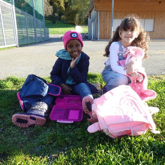 Zwei junge Mädchen sitzen auf Gras vor einem Gebäude. Ein Mädchen trägt einen rosa Hut und Rucksack. Beide Mädchen haben Schultaschen in der Nähe.