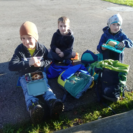 Drei Kinder sitzen auf der Straße, jedes mit einer Brotdose und einem Rucksack. Das Kind auf der linken Seite lächelt, während die anderen ernst aussehen. Der Junge auf der rechten Seite trägt eine Mütze und eine Schürze.