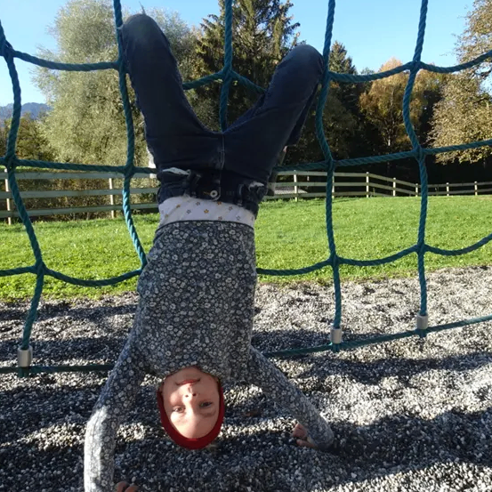 Ein Kind hängt kopfüber an einem blauen Kletternetz in einem Spielplatz, mit Kies darunter und einem Grasfeld im Hintergrund.