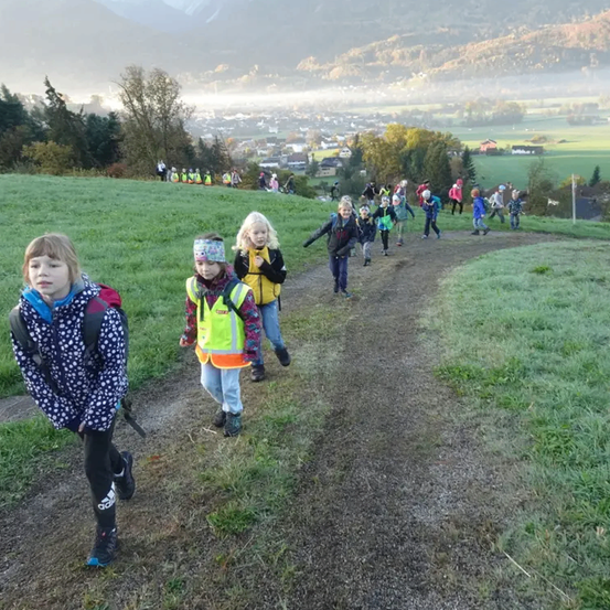 Eine Gruppe von Kindern, einige in gelben Westen, gehen auf einem Feldweg in einem Grasfeld, mit Bergen und einem Dorf im Hintergrund.