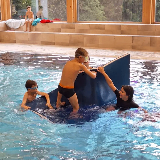 Zwei junge Jungen und eine Frau sind in einem Innenpool. Ein Junge hält ein blaues Objekt, und der andere steht darauf. Die Frau hilft dem Jungen. Ein Junge sitzt auf einer Bank in der Nähe eines Fensters mit Bäumen draußen.