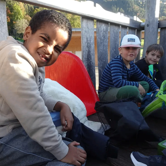 Drei Kinder sitzen auf einer Bank. Ein Junge lächelt und schaut in die Kamera. Ein anderer trägt einen Baseball-Cap. Der dritte Junge trägt einen grünen Mantel.