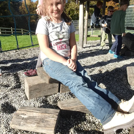 Ein kleines Mädchen sitzt auf einem Stapel hölzerner Baumstämme in einem Spielplatz im Freien. Ein anderes Kind steht hinter ihr, und mehrere andere Kinder sind in der Nähe. Die Umgebung ist draußen, mit einem klaren Himmel und Bäumen im Hintergrund.