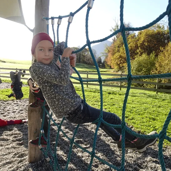 Ein kleines Kind klettert in einem Park an einem grünen Seilnetz und trägt einen roten Hut und Turnschuhe. Ein anderes Kind ist am Boden.