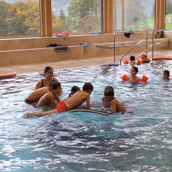 Mehrere Kinder schwimmen in einem Pool. Sie tragen Schwimmbrillen und Schwimmwesten. Eine erwachsene Frau schwimmt hinter ihnen. Der Pool hat Glaswände.
