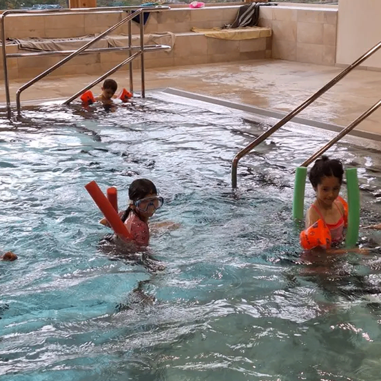 Kinder schwimmen in einem Pool mit Schwimmbrillen und Schwimmhilfen, eins hält einen roten Schwimmstab, umgeben von Handläufen und einer Bank mit einer Decke.