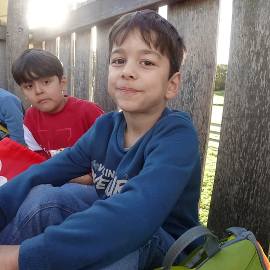 Zwei Jungen sitzen auf einem Holzzaun. Ein Junge trägt ein blaues Sweatshirt und der andere ein rotes Shirt. Beide haben Schulranzen in der Nähe.