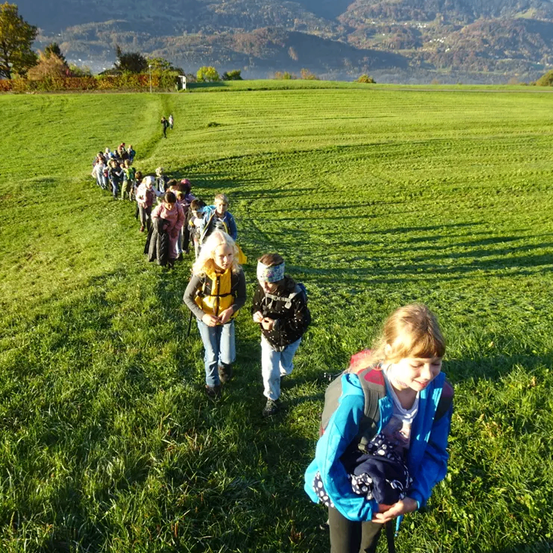Eine Gruppe von Kindern geht auf einem Feld, eine hält ein Baby. Sie sind in einer Reihe mit einer Person, die führt. In der Ferne gibt es eine Bergkette.