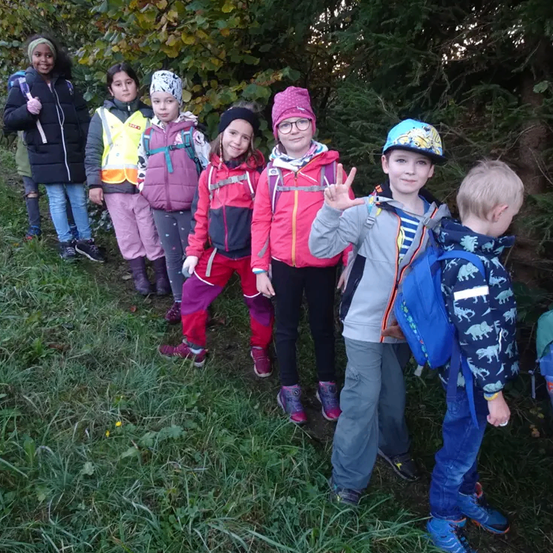Eine Gruppe von Kindern steht auf einer Rasenfläche. Einige tragen Jacken und Mützen. Ein Junge macht ein Friedenszeichen. Hinter ihnen stehen Bäume und Büsche.
