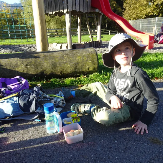 Ein Junge sitzt lächelnd auf dem Boden und trägt einen Hut. In der Nähe befinden sich ein Brotkasten, eine Flasche und ein Rucksack. Hinter ihm sind ein roter Rutschen und ein Spielhaus zu sehen.