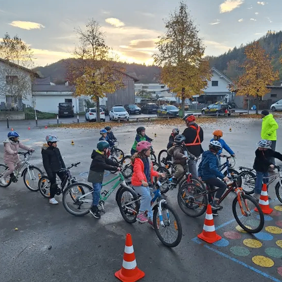 Eine Gruppe von Kindern fährt auf einem Parkplatz mit Fahrrädern, angeführt von einer Person in einer gelben Jacke. Die Fahrräder sind in einem Kreis mit um sie herum aufgestellten Verkehrskegel angeordnet. Dahinter sind Autos geparkt, und Gebäude sind sichtbar. Bäume sind ebenfalls vorhanden, und der Himmel ist bewölkt.