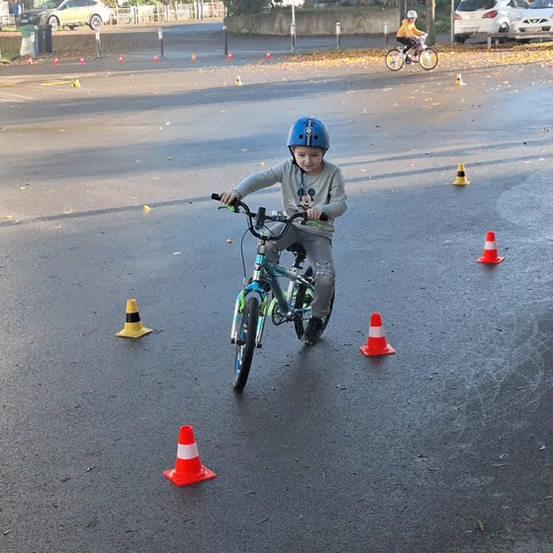 Ein junges Kind fährt mit einem Fahrrad zwischen orangefarbenen Hütchen auf einer leeren Straße und trägt einen Helm. Ein weiterer Fahrer folgt dahinter. Bäume und geparkte Autos sind im Hintergrund zu sehen.