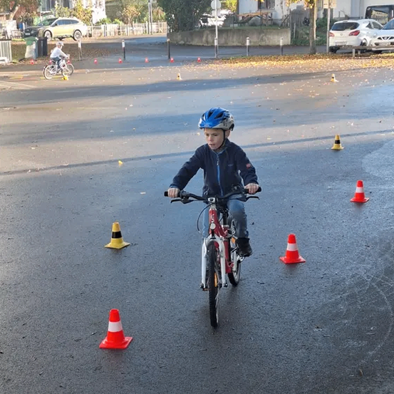 Ein Junge fährt auf einer nassen Straße mit einem Fahrrad und navigiert durch Verkehrskegel. Im Hintergrund fährt ein weiteres Kind ein Fahrrad, und es gibt Häuser, Autos und Bäume.