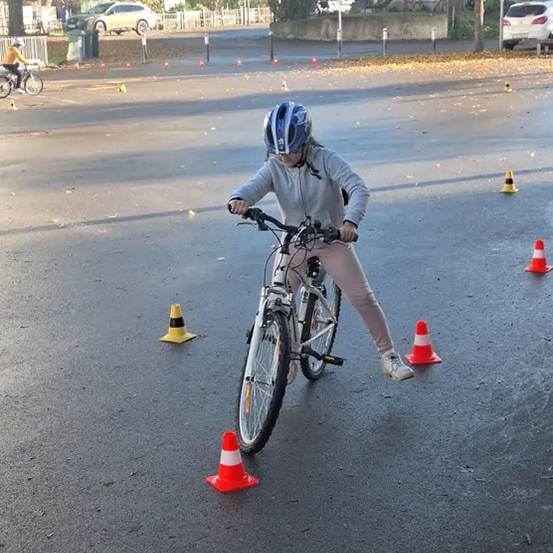 Ein junges Mädchen fährt ein Fahrrad auf einer Straße, umgeben von orangefarbenen und weißen Hütchen. Sie trägt einen Helm und Turnschuhe. Hinter ihr fährt ein weiterer Radfahrer in der Ferne.