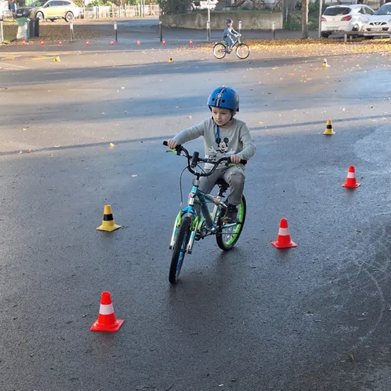 Ein Kind fährt ein Fahrrad zwischen orangefarbenen Verkehrskegel auf einer nassen Straße. Ein kleiner Erwachsener fährt im Hintergrund ein Fahrrad.