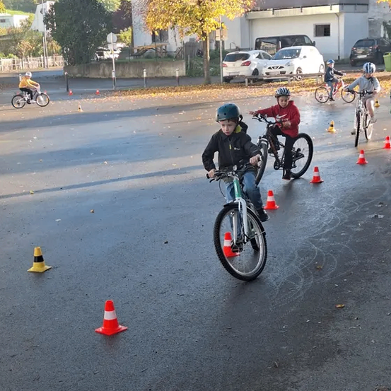 Mehrere Kinder fahren auf einer nassen Straße mit Fahrrädern und tragen Helme. Sie navigieren durch orange und gelbe Verkehrskegel. Im Hintergrund sind geparkte Autos und ein Gebäude mit einer Veranda zu sehen.
