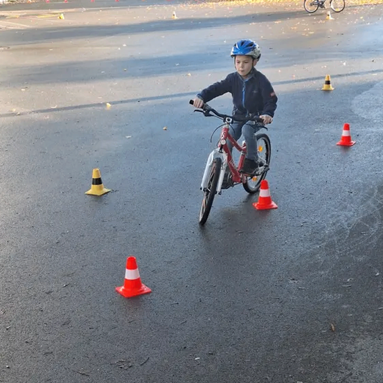 Ein Kind mit Helm fährt auf einer nassen Straße mit dem Fahrrad und navigiert durch Verkehrskegel. Im Hintergrund fährt ein weiteres Kind mit dem Fahrrad, und Autos sind in der Nähe eines Gebäudes geparkt.