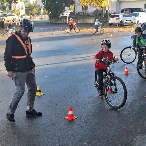 Eine Gruppe von Kindern fährt auf einem Parkplatz mit Fahrrädern. Sie tragen Helme und es gibt orangefarbene Verkehrskegel um sie herum. Ein Erwachsener geht neben einem Jungen her, der ein Fahrrad fährt.