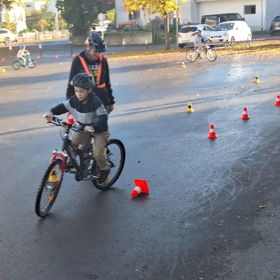 Ein Junge fährt sein Fahrrad durch eine Hütchenbahn, begleitet von einem Mann in Sicherheitsausrüstung. Mehrere Kinder fahren im Hintergrund Fahrräder.