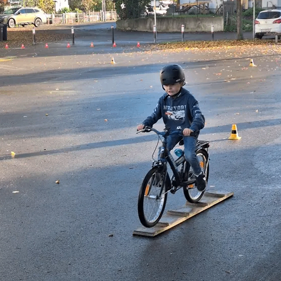 Ein junger Junge fährt auf einem Parkplatz auf einer Rampe mit einem Fahrrad, trägt einen Helm und Jeans. Es gibt Verkehrskegel und geparkte Autos in der Nähe.