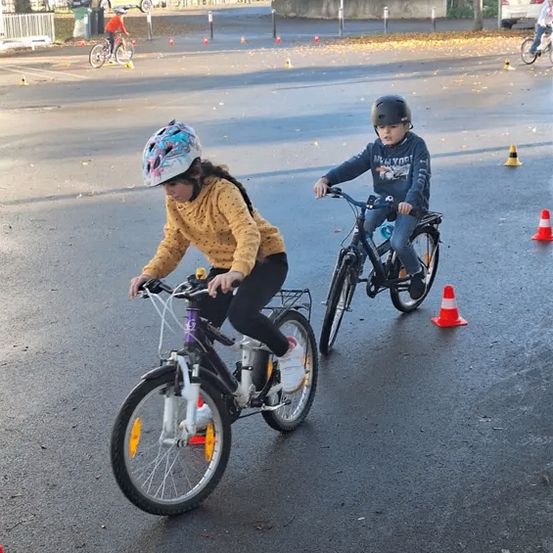 Zwei Kinder fahren Fahrräder auf einer Straße, die mit Verkehrskegeln gesäumt ist. Sie tragen Helme und Jacken. Dahinter sind mehrere Fahrräder und Autos geparkt. Bäume und Gebäude säumen die Straße.