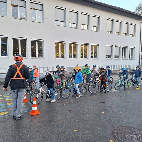 Eine Gruppe von Kindern in Helmen übt das Radfahren auf einem Parkplatz. Ein Mann in einer reflektierenden Weste beaufsichtigt sie. Dahinter befindet sich ein Gebäude mit vielen Fenstern.