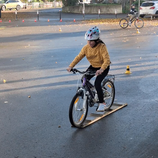 Ein junges Mädchen fährt mit einem Fahrrad über eine Rampe auf einer leeren Straße mit Verkehrskegeln und geparkten Autos.