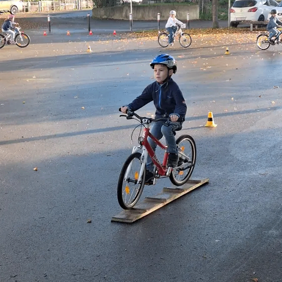 Ein Junge fährt auf einer asphaltierten Straße mit dem Fahrrad, in der Nähe reiten mehrere Kinder Fahrräder. Der Junge trägt einen Helm. Die Straße ist mit Verkehrskegeln markiert.