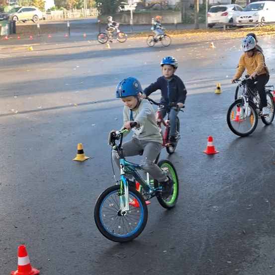 Kinder, die Helme tragen, fahren mit Fahrrädern auf einer nassen Straße mit Kegel. Mehrere Autos sind in der Nähe geparkt.