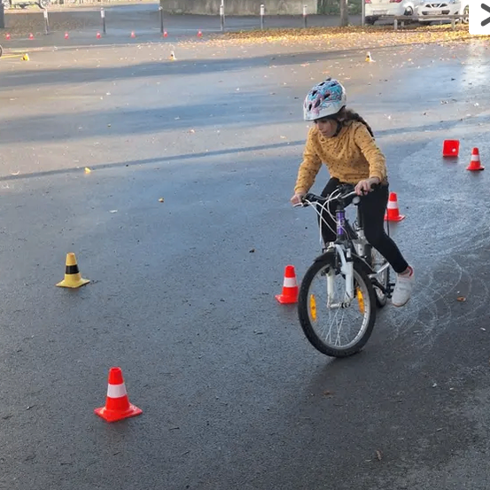 Ein junges Mädchen fährt mit einem Fahrrad durch eine nasse Straße mit aufgestellten Verkehrskegeln. Sie trägt einen Helm und Turnschuhe. Die Straße hat geparkte Autos und Gebäude im Hintergrund.