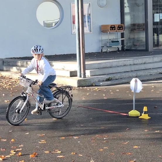 Ein junges Kind fährt mit einem Fahrrad um Kegel auf einer gepflasterten Oberfläche und trägt einen Helm. Dahinter führt ein Gebäude mit einer Glastür und Stufen zu einem Weg.