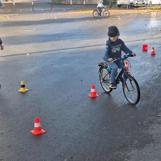 Ein junger Junge fährt auf einer nassen Straße mit einem Fahrrad und navigiert um orangefarbene Verkehrskegel herum. Ein anderes Kind fährt im Hintergrund ein Fahrrad. Mehrere Autos sind am Straßenrand geparkt.