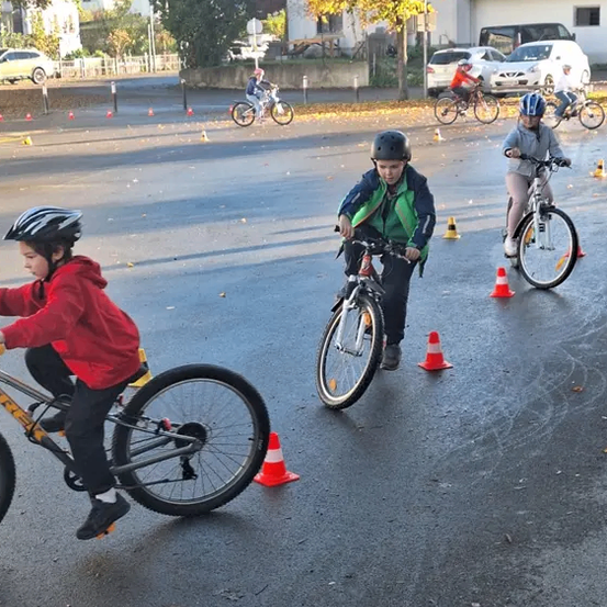 Eine Gruppe von Kindern fährt auf einer nassen Straße mit Fahrrädern und navigiert um Verkehrskegel herum, mit Bäumen und Gebäuden im Hintergrund.