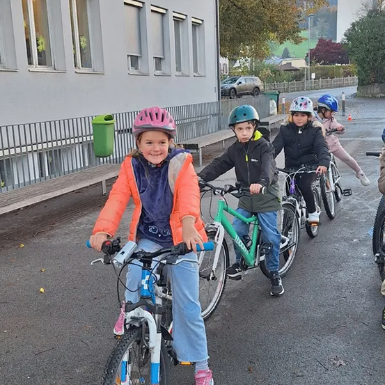 Eine Gruppe von Kindern fährt auf einer Straße mit Fahrrädern, mit einem Gebäude und Bänken im Hintergrund.