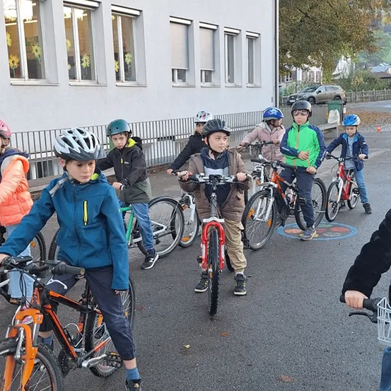 Mehrere Kinder, die Helme tragen, fahren in einer Reihe vor einem Gebäude mit großen Fenstern mit Fahrrädern. Ein orangefarbener Verkehrskegel befindet sich im Vordergrund.
