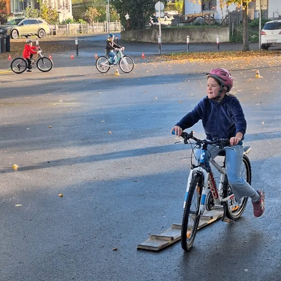 Ein Kind fährt auf einer Straße mit dem Fahrrad. In der Nähe sind andere Kinder, die Fahrräder fahren. Sie befinden sich in einem Trainingsbereich mit Hütchen und einer Rampe. Im Hintergrund sind Häuser und Autos zu sehen.