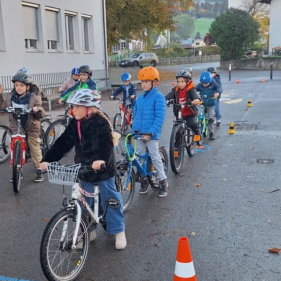 Eine Gruppe von Kindern fährt mit Fahrrädern auf einem Parkplatz, mit einem Gebäude und Bäumen im Hintergrund.