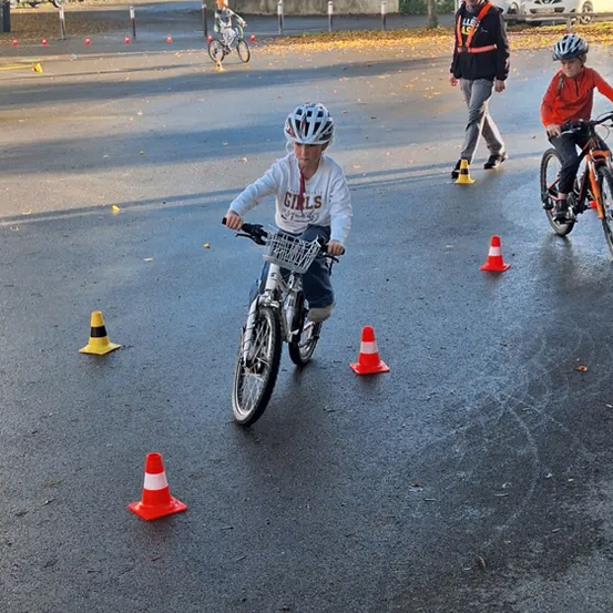 Kinder fahren mit Fahrrädern durch einen Parcours, der mit Hütchen markiert ist. Ein Kind trägt ein Shirt mit der Aufschrift 'GIRLS'. Ein anderes Kind trägt einen Helm und hat einen Korb am Fahrrad.