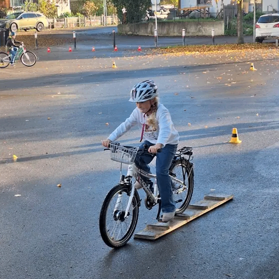 Ein Kind fährt mit einem Fahrrad auf einer Rampe in einem leeren Parkplatz mit orangefarbenen Kegeln drumherum.