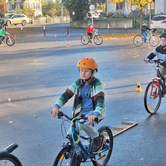 Mehrere Kinder fahren mit Fahrrädern in einem Parkplatz mit kleinen Verkehrskegeln und Bäumen im Hintergrund.