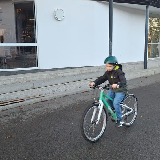 Ein junges Kind mit einem grünen Helm fährt ein grünes Fahrrad auf einer asphaltierten Straße vor einem weißen Gebäude mit Glasfenstern.