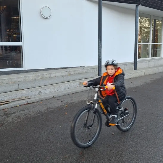 Ein Kind fährt ein Fahrrad auf einer asphaltierten Straße vor einem weißen Gebäude mit Glasfenstern.