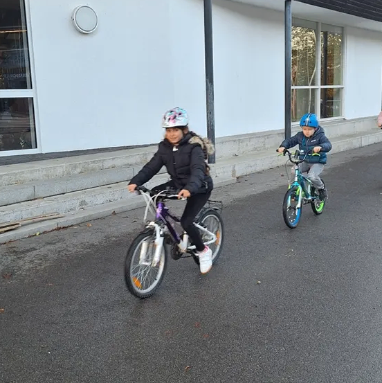 Zwei Kinder fahren auf einem asphaltierten Weg vor einem Gebäude mit großen Fenstern mit Fahrrädern. Beide tragen Helme und Jacken.