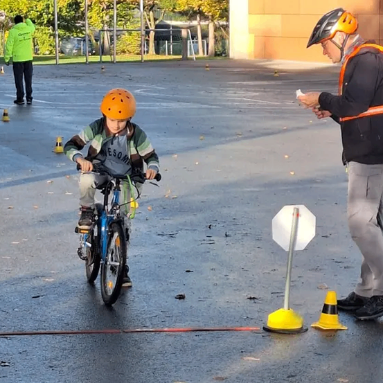 Ein junger Junge fährt mit einem Helm auf einer nassen Straße mit dem Fahrrad. Ein Mann mit einer reflektierenden Weste gibt Anweisungen. Im Hintergrund befinden sich Verkehrskegel und Stangen.