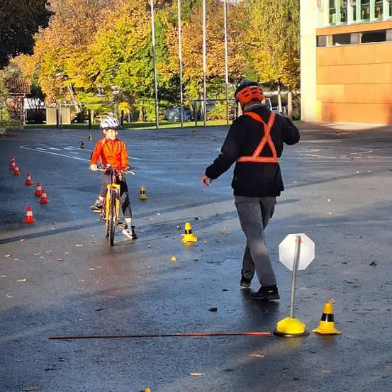 Ein Kind fährt mit dem Fahrrad durch einen Hindernisparcours auf einem Parkplatz, wobei ein Erwachsener in der Nähe ist.